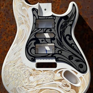 May include: A white electric guitar body with intricate floral designs carved into the wood. The body is painted white and the designs are in a light brown color. The guitar is a blank canvas for a custom paint job.