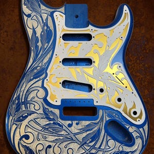 May include: A blue electric guitar body with a gold and silver engraved floral design. The design features intricate leaves and swirls. The text "DUSTSTORM ENGRAVING" is visible at the top of the image.