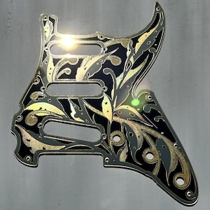 May include: A black and gold guitar pickguard with an ornate floral design. The pickguard has three pickup cutouts and is made in the USA. The text "Dust Storm Engraving" is at the bottom.