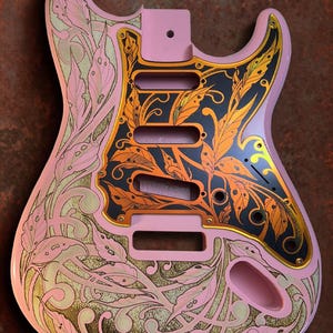 May include: A pink electric guitar body with a black and gold leaf design. The design is engraved into the body and features a floral pattern. The guitar body is blank, with no pickups or other hardware.