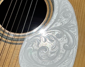 May include: A clear, pear-shaped guitar pickguard with an engraved floral design, set against the wood grain of an acoustic guitar. The pickguard is positioned near the sound hole and strings. The text "DUST STORM ENGRAVING" is visible at the bottom.