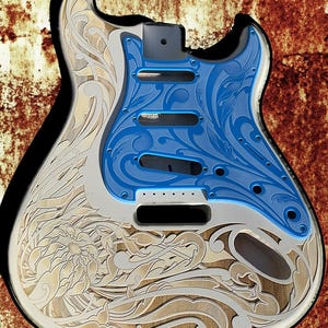 Op de afbeelding: Een witte elektrische gitaar body met een blauw en wit bloemenpatroon. De gitaar body is gegraveerd met een gedetailleerd bloemenpatroon. De tekst "DUSTSTORM ENGRAVING" is zichtbaar bovenaan de afbeelding.