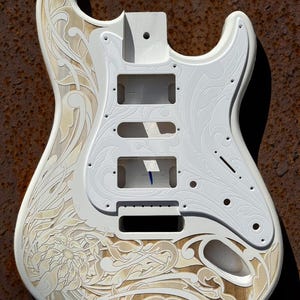 May include: White electric guitar body with intricate floral and scrollwork engraving. The body is made of wood and has a natural wood grain finish. The engraving is done in a light brown colour. The guitar body is ready for painting or finishing.