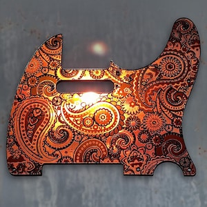 Op de afbeelding: Een koperkleurige gitaar pickguard met een paisley-patroon. De pickguard is gemaakt van metaal en heeft een glanzende afwerking.