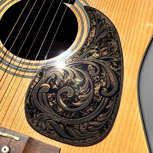 May include: Close-up of an acoustic guitar with a decorative black and gold pickguard featuring an ornate, swirling design. The guitar's wooden body has visible grain, and the strings are taut. The pickguard is from Duststorm Engravings.