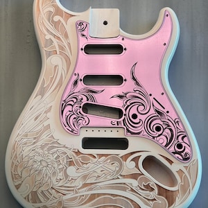 May include: A white electric guitar body with a pink pickguard. The body is decorated with an intricate floral design carved into the wood. The pickguard also has a floral design, but it is black and white. The text "DUSTSTORM ENGRAVING" is visible in the top right corner of the image.