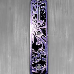 May include: A black and purple engraved metal plate with a floral design. The plate has a rectangular shape with rounded ends and several holes. The text "DUST STORM ENGRAVING" is at the top left.