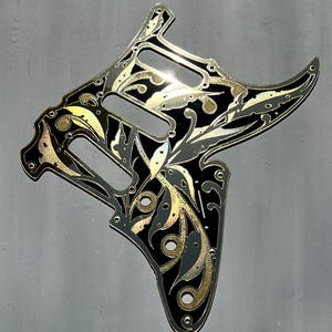 May include: Black and gold guitar pickguard with an ornate floral design. The pickguard has cutouts for the pickups and controls. The words "Dust Storm Engraving" are at the bottom. The pickguard is made in the USA.