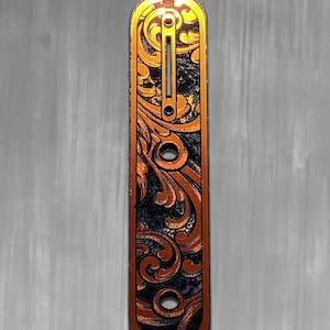 May include: A decorative, elongated metal piece with an intricate, swirling floral design. The piece features a black background with copper-colored accents and a gold border. The top left corner displays the text "DUST STORM ENGRAVING".