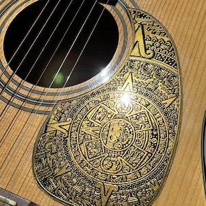 May include: Acoustic guitar with a custom pickguard featuring a gold Aztec calendar design. The pickguard is intricately detailed and contrasts with the natural wood grain of the guitar. The text "DUST STORM ENGRAVING" is visible at the bottom.