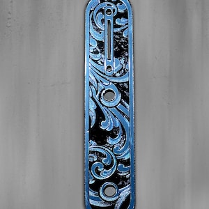May include: A blue and black engraved metal plate with a decorative floral design. The plate has a rectangular shape with rounded ends and several holes. The text "DUST STORM ENGRAVING" is at the top left.