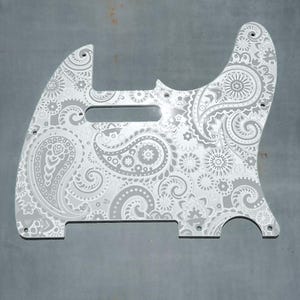 Op de afbeelding: Een witte gitaar pickguard met een paisley-patroon. De pickguard is gemaakt van een duurzaam materiaal en is ontworpen om te passen op de meeste standaard elektrische gitaren.