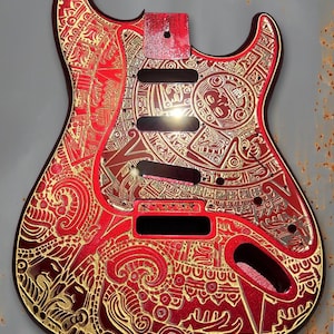 Peut inclure: Corps de guitare rouge et or avec des motifs gravés complexes. Le corps présente une finition rouge vif avec des motifs dorés détaillés, notamment des motifs géométriques et stylisés. Le texte "Dust Storm Engraving" est visible.