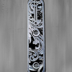 May include: A silver-colored, elongated metal plate with an ornate, engraved floral design. The plate has a black background and several circular holes. The top left corner has the text "DUST STORM ENGRAVING". The American flag is in the top right corner.