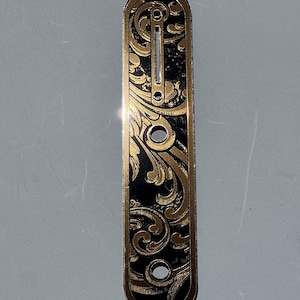 May include: A black and gold engraved metal plate with an ornate floral design. The plate has a rectangular shape with rounded edges and several holes. The text "DUST STORM ENGRAVING" is visible in the upper left corner.