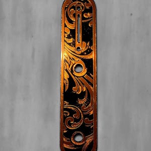 May include: A black and copper guitar part with an ornate floral design. The part is rectangular with rounded ends and several holes. The text "DUST STORM ENGRAVING" is at the top left.
