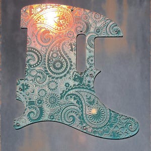 Op de afbeelding: Een teal en koperkleurige gitaar slagplaat met een ingewikkeld paisley-ontwerp. De slagplaat is gegraveerd met bloemen- en wervelpatronen. De tekst "DUST STORM ENGRAVING" staat onderaan, en "*OUTDOOR DAYLIGHT" eronder. De afbeelding bevat ook een "PROUDLY MADE IN USA" sticker.