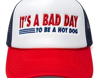 Könnte beinhalten: Eine Trucker-Cap mit weißer Schaumstofffront, rotem Schirm und dunkelblauem Netzrücken. Auf der Vorderseite befindet sich der rote Stickschriftzug IT'S A BAD DAY und der blaue Schriftzug TO BE A HOT DOG mit drei horizontalen blauen Linien.