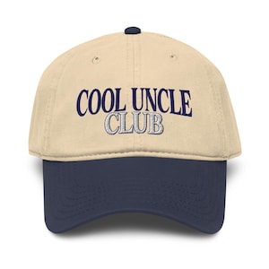 May include: A beige baseball cap with a navy blue brim. The cap features the embroidered text "COOL UNCLE CLUB" in navy blue and white. The cap has a curved brim and a button on top.