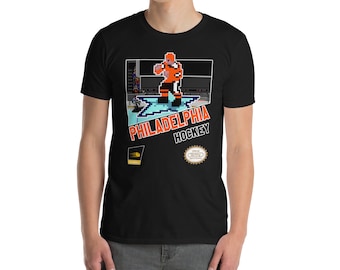 Puede incluir: Camiseta negra con un diseño de videojuego retro que presenta a un jugador de hockey sobre el hielo. El diseño incluye el texto "Philadelphia Hockey" y un logotipo de Nintendo Entertainment System.