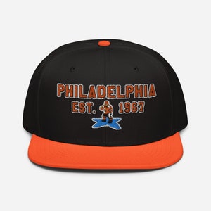 May include: Black and orange baseball cap with the text "PHILADELPHIA EST. 1967" and a cartoon figure in the center.