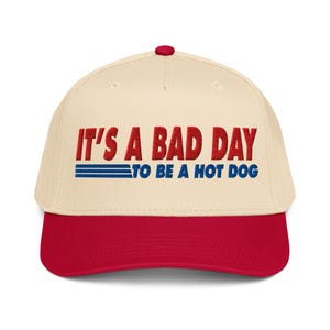 May include: A baseball cap with a cream-colored crown and a red brim. The cap features the text "IT'S A BAD DAY TO BE A HOT DOG" in red and blue lettering. The cap has a red button on top.