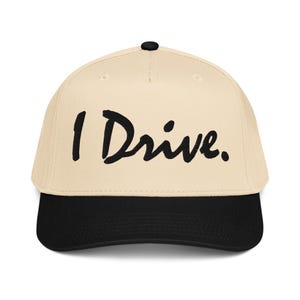 May include: A baseball cap with a cream-colored crown and a black brim. The phrase "I Drive." is printed in black, cursive-style lettering across the front of the cap. The cap has a black button on top.