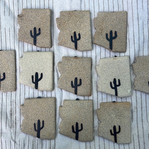 Può includere: Una collezione di piastrelle in ceramica a forma di Arizona in varie tonalità di beige e marrone. Ogni piastrella presenta un disegno di cactus nero. Le piastrelle sono disposte su un tessuto a righe bianco sporco.