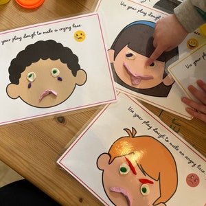 May include: Three white cards with cartoon faces on them. Each card has instructions to use play dough to make a different facial expression: crying, smiling, and angry. The cards are on a wooden table.
