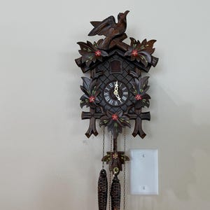 May include: A dark brown wooden cuckoo clock featuring a carved bird at the top, intricate leaf motifs with small red accents, and a circular clock face with Roman numerals. Two dark pinecone-shaped weights hang from chains below the clock body.