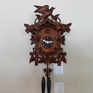 May include: A dark brown wooden cuckoo clock featuring intricate carved leaf details and a bird perched at the top. The clock face is circular with Roman numerals and white hands. Two pinecone-shaped weights hang from chains at the bottom.