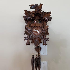 May include: A dark brown wooden cuckoo clock featuring intricate carved leaf motifs, a bird perched at the top, and two small figures in a balcony above the clock face. Three pinecone-shaped weights hang from chains at the bottom of the clock.