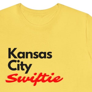 Puede incluir: Camiseta amarilla con texto negro que dice "Kansas City" y texto rojo que dice "Swiftie".