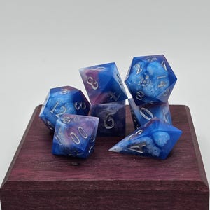 Puede incluir: Un conjunto de siete dados poliédricos en tonos azules, morados y blancos, con números plateados. Los dados se exhiben sobre un bloque de madera morada oscura. Para juegos de mesa.