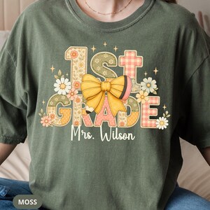 Könnte beinhalten: Ein moosgrünes T-Shirt mit einem Aufdruck, der den Text 1st GRADE und Mrs. Wilson zeigt. Das Design enthält eine große gelbe Schleife, einen Bleistift sowie verschiedene Blumen- und Vichy-Karomuster in Pastelltönen, akzentuiert durch kleine Sterne.