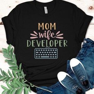 Puede incluir: Camiseta negra con el texto "MOM wife DEVELOPER" en letras de colores y un gráfico de teclado.