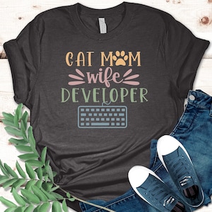 May include: Dark gray t-shirt with the text "Cat Mom Wife Developer" in orange, pink, and green. A light blue keyboard graphic is below the text. The shirt is paired with blue jeans and navy blue sneakers.
