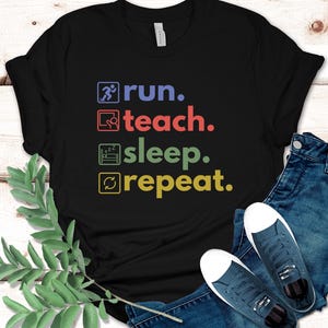Puede incluir: Camiseta negra con un gráfico que dice "run. teach. sleep. repeat." en cuadros de colores.