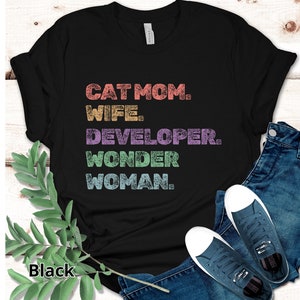 May include: Black t-shirt with the words "CAT MOM. WIFE. DEVELOPER. WONDER WOMAN." in colorful, distressed text. The shirt is paired with blue jeans and navy blue sneakers.