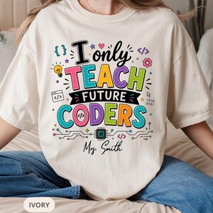 Peut inclure: Un t-shirt ivoire avec un motif coloré portant le texte "I only TEACH FUTURE CODERS" et "Mrs. Smith". Le design comprend des icônes comme une ampoule, une puce informatique, du code binaire et des crochets en rose, jaune, violet et bleu.