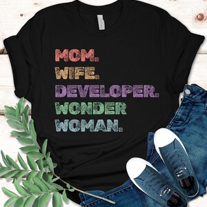May include: Black t-shirt with the words "MOM. WIFE. DEVELOPER. WONDER WOMAN." in colorful, distressed text. The shirt is paired with blue jeans and navy blue sneakers.