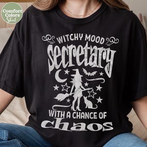 Pode incluir: Camiseta preta com um gráfico branco que diz "Witchy Mood Secretary With a Chance of Chaos". O design inclui uma silhueta de bruxa, estrelas, uma lua crescente, um morcego e um gato. Marca Comfort Colors.