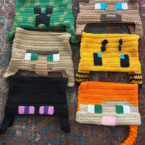 May include: A collection of six crocheted hats, each designed to resemble characters from a popular video game. The hats feature various colors, including green, brown, yellow, black, and orange, with pixelated facial features and unique designs.