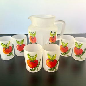 May include: A white pitcher and six matching glasses. Each piece is decorated with a hand-painted design of a red tomato with green leaves. The pitcher has a handle and a spout. The glasses are cylindrical.