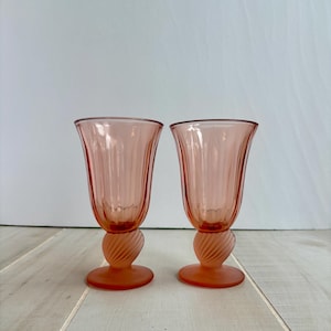 May include: Two pink-tinted glass goblets. The goblets have a fluted design on the bowl and a twisted stem. The base is a flat, circular design. The goblets are displayed on a light-colored wooden surface.