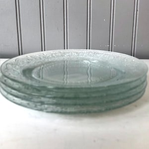 May include: A stack of four clear glass plates with a decorative embossed pattern around the edge.