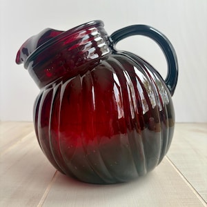 May include: A red glass pitcher with a ribbed design and a black handle. The pitcher is sitting on a light wood surface.