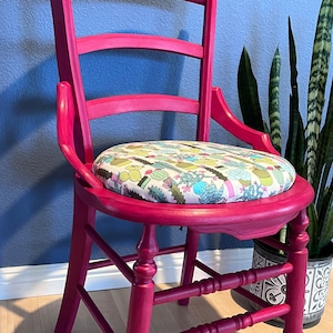 May include: A bright pink wooden chair with a floral patterned cushion. The chair has a ladder back and turned legs.