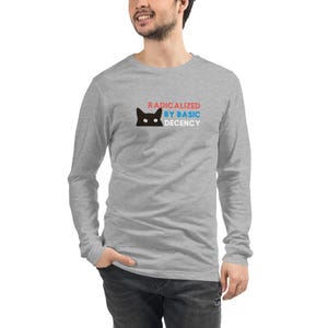 Könnte beinhalten: Ein langärmeliges Shirt in Grau meliert mit einer Grafik eines schwarzen Katzenkopfes mit weißen Augen. Der Text RADICALIZED ist in Rot, BY BASIC in Blau und DECENCY in Weiß gedruckt und befindet sich rechts neben der Katzengrafik auf der Brust.