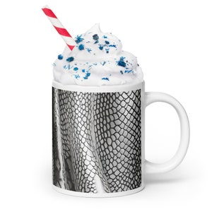 Könnte beinhalten: Weiße Keramiktasse mit einem schwarz-weißen Schlangenhautmuster. Die Tasse hat einen weißen Rand und einen weißen Henkel. Die Tasse ist mit Schlagsahne und blauen Streuseln gefüllt. Ein rot-weiß gestreifter Strohhalm ragt aus der Schlagsahne.